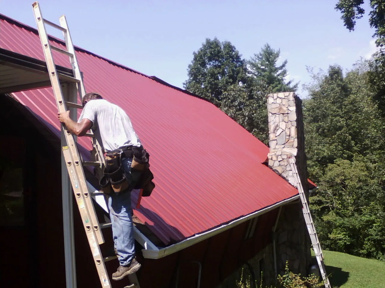 Roofer on a ladder beside a red metal roof and stone chimney — Soffit & Fascia Repair by Roofing Brothers in Melvindale